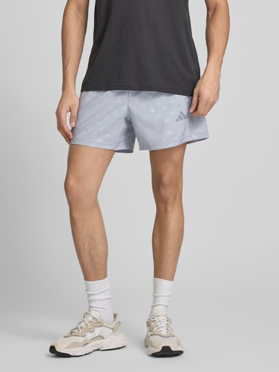 Mężczyzna nosi Regular Fit szorty z logo na całej powierzchni model 'WE BRLO SHO' od adidas Training, efekt: jednolity kolor, wykończenie materiału: satyna, detal z logo, elastyczny ściągacz, boczne kieszenie zapinane na zamek błyskawiczny, krój regular fit, wzór na całej powierzchni, widok z przodu na całe ciało.