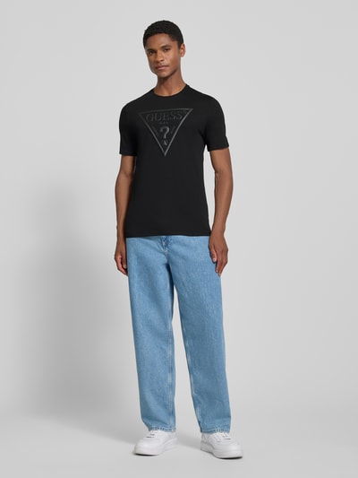 Männermodel trägt T-Shirt mit Logo und Rundhalsausschnitt von GUESS in Schwarz, Reine Baumwolle, Slim-Fit, Rückenlänge 70 cm, Ganzkörperansicht von vorne.