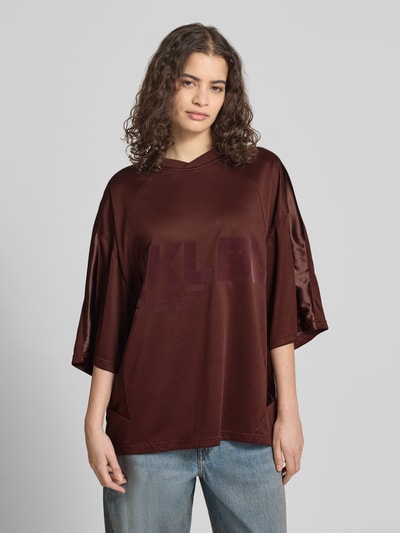 Kobiece model prezentuje Oversized T-Shirt mit Label-Print von Karo Kauer, damski t-shirt o kroju oversized, nadruk z logo, dekolt w serek, opuszczone ramiona, długość tyłu w rozmiarze S: 71 cm, ciemnobrązowy, gładka faktura, całkowity widok od przodu