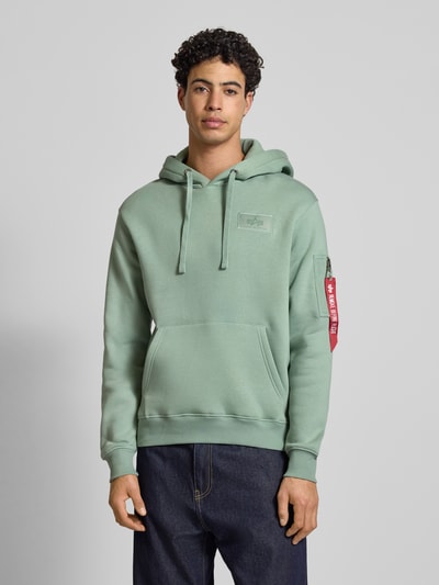 Männermodel trägt Hoodie mit Känguru-Tasche von Alpha Industries, Regular-Fit, Baumwoll-Mix, Label-Patch, gerade Farben, Nahaufnahme Oberkörper.