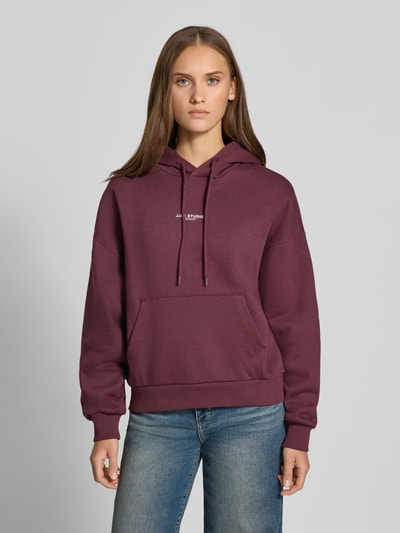 Frauenmodel trägt Relaxed Fit Hoodie aus Baumwoll-Mix Modell 'VESTERBRO' von JJXX in Dunkelrot mit Label-Detail, Känguru-Tasche, Kapuze mit Tunnelzug, Ganzkörperansicht von vorne.