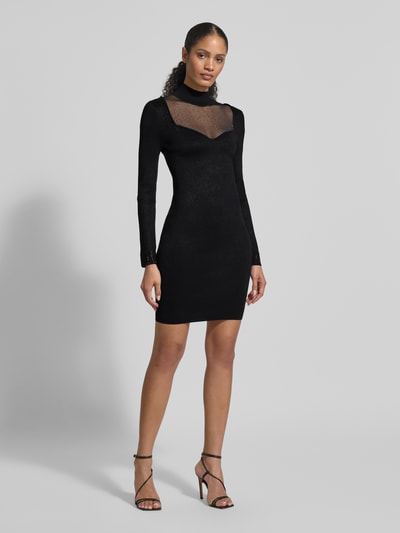 Frauenmodel trägt Slim Fit Kleid aus Viskose-Mix mit Ziersteinbesatz Modell 'Lucy' von Guess, schwarz, semitransparent, Langarm, Stehkragen, Ganzkörperansicht von vorne.