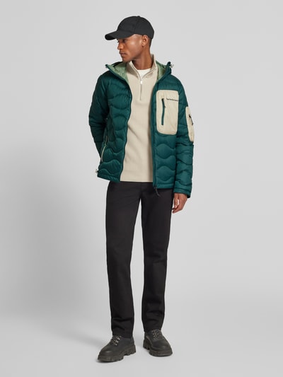 Männermodel trägt Daunenjacke mit Kapuze von PEAK PERFORMANCE in Dunkelgrün mit Reißverschlüssen und Brusttasche, Ganzkörperansicht von vorne. Gerader Schnitt, Kapuze, Tunnelzug, Reißverschlusstaschen, Rückenlänge 73 cm, Schulterbreite 49 cm, Brustweite 58 cm, Ärmellänge 67 cm.