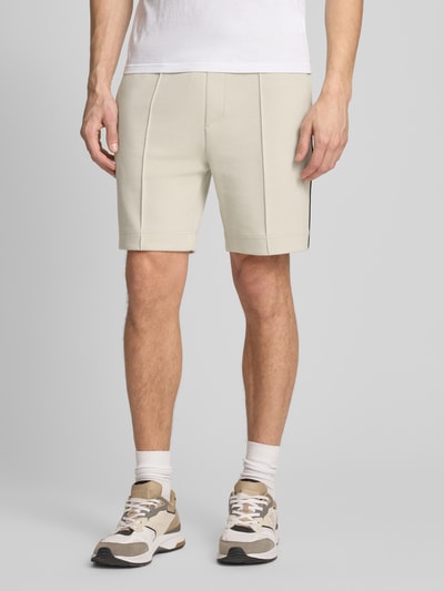 Männermodel trägt beigefarbene Shorts mit glatter Twill-Optik und gerader Form, Kontraststreifen an der Seite, Ganzkörperansicht von vorne.