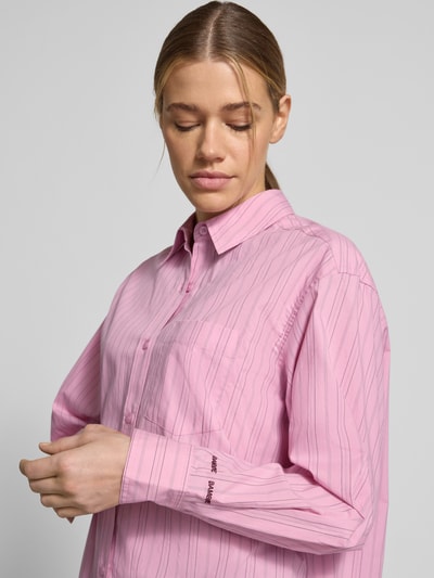 Modelka nosi Regular Fit Bluse mit Brusttasche Modell 'LOVA' od Samsøe Samsøe, czysta bawełna, krój regular fit, kołnierzyk koszulowy, kieszeń na piersi, haft z logo, długość tyłu w rozm. S 74 cm, długość rękawa 59 cm, różowe paski, miękka tekstura, zbliżenie na górną część ciała z przodu.