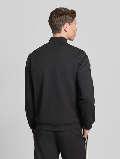 Männermodel trägt Sweatjacke mit Stehkragen und Zweiwege-Reißverschluss von EA7 Emporio Armani, czarny kolor, krój regular fit, detal z logo, widok całej sylwetki z tyłu.