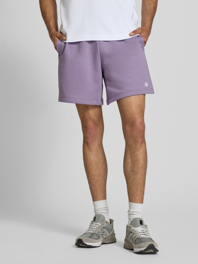 Männermodel trägt Relaxed Fit Shorts mit Logo-Stitching von New Balance in Lila, Baumwoll-Mix, elastischer Bund mit Tunnelzug, Ganzkörperansicht von vorne.