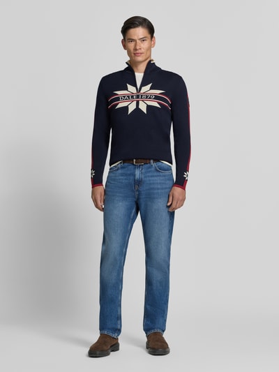 Männermodel trägt Wollpullover mit Label-Print Modell 'Nale' von Dale of Norway in Dunkelblau mit roten Streifen, Ganzkörperansicht von vorne, Reine Wolle, gerader Schnitt, Troyer-Kragen, Rückenlänge 67 cm, Ärmellänge 68 cm.