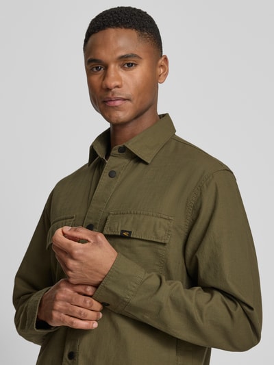 Männermodel trägt Regular Fit Hemdjacke aus reiner Baumwolle von camel active in Olivgrün mit Durchgehender Knopfleiste, Statement-Patch, Label-Detail, Brusttaschen, Langärmelig, Nahaufnahme Oberkörper.