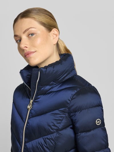 Frauenmodel trägt Steppmantel mit Kapuze und Reißverschlusstaschen von MICHAEL Michael Kors in Dunkelblau, glänzende Textur, Nahaufnahme Oberkörper. Gerader Schnitt, Stehkragen, seitliche Reißverschlusstaschen, sichtbares Markenlogo auf dem Ärmel.