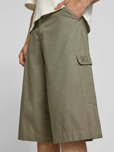 Męski model nosi Straight Fit Shorts mit Cargo-Taschen Modell 'Camo' od KARL KANI z bawełny, kieszenie cargo, wpuszczane i z tyłu, zapięcie na guzik i zamek, szerokość w rozmiarze M: 41 cm, długość wewnętrzna nogawki: 38 cm, jednolity oliwkowy kolor, prosty krój, całkowity widok z przodu