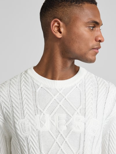 Männermodel trägt weißen Strickpullover aus Baumwoll-Modal-Mix von Guess, Zopfmuster, Rundhalsausschnitt, Nahaufnahme Oberkörper.