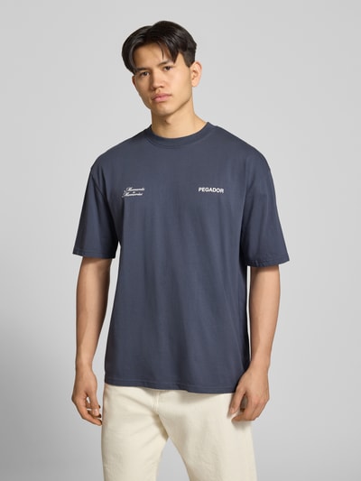 Męski model nosi Oversized T-Shirt mit Label-Detail Modell 'Rochefort' von Pegador, czysta bawełna, okrągły dekolt, nadruk z tyłu, kolor ciemnoszary, widok całej sylwetki z przodu.