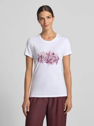 Kobiece model prezentuje Damski t-shirt Regular Fit T-Shirt aus reiner Baumwolle von Liu Jo White, bawełna, nadruk z logo, obszycie z kamieni stras, okrągły dekolt, długość tyłu w rozmiarze S: 66 cm, widok całej sylwetki z przodu.