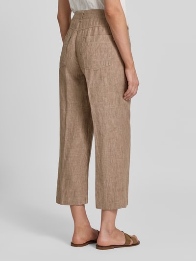 Frauenmodel trägt kuloty z lnu z wpuszczanymi kieszeniami model 'Nora' od MAC, krój wide leg fit, detal z logo, zapięcie na guzik i zamek błyskawiczny, szerokość w rozmiarze 36: 44 cm, długość wewnętrzna nogawki w rozmiarze 36: 67 cm, kolor jasnobrązowy, widoczne wyraźne faktury lnu, całkowity widok z tyłu.