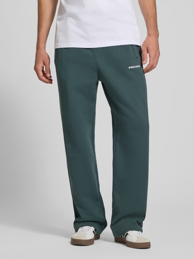 Männermodel trägt Loose Fit Sweatpants aus Baumwoll-Mix mit Label-Detail von PEGADOR, grün, mit Eingrifftaschen, Ganzkörperansicht von vorne.