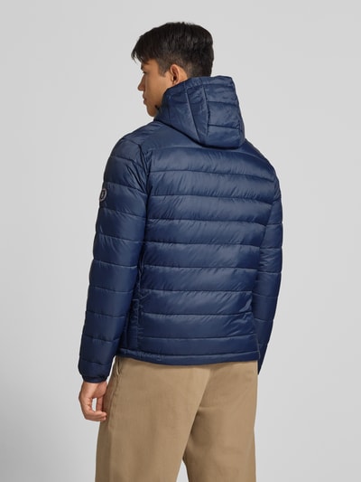 Męski model nosi Regular Fit kurtkę w stylu puffer z efektem pikowania Model 'KINGSLEY' od JACK & JONES, kolor granatowy, kaptur, zamek błyskawiczny, wpuszczane kieszenie, długość tyłu w rozmiarze M: 70 cm, szerokość ramion w rozmiarze M: 45 cm, szerokość w rozmiarze M: 52 cm, długość rękawów w rozmiarze M: 67 cm, zdjęcie w całej sylwetce z tyłu.