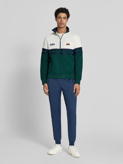 Männermodel trägt Sweatjacke aus Baumwoll-Mix Modell 'RIMINI' von Ellesse, Farbkombination Weiß, Grün und Navy, Gerade Passform, Reißverschluss, Stehkragen, Label-Details, elastische Abschlüsse, Ganzkörperansicht von vorne.