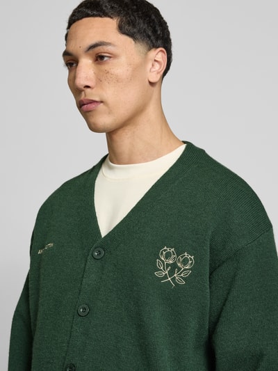 Männermodel trägt Cardigan mit Logo-Stitchings und V-Ausschnitt von ANOTHER COTTON LAB, grün, grobmaschige Textur, Nahaufnahme Oberkörper.