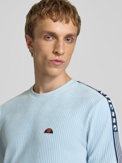 Męski model nosi T-Shirt mit Logo Modell 'MADORI' od Ellesse z mieszanki bawełny, krój regular fit, okrągły dekolt, logo, długość tyłu 66 cm w rozmiarze M, jasnoniebieski, prążkowana faktura. Nahaufnahme górnej części ciała.