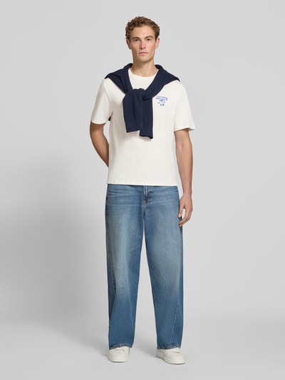 Männermodel trägt Regular Fit T-Shirt aus reiner Baumwolle Modell 'VIBING COCO' von REVIEW in Weiß, hellblaue Jeans, Ganzkörperansicht von vorne, Herren T-Shirt von REVIEW x ANSON´S, Print auf der Rückseite, Rückenlänge bei Größe M: 68 cm.