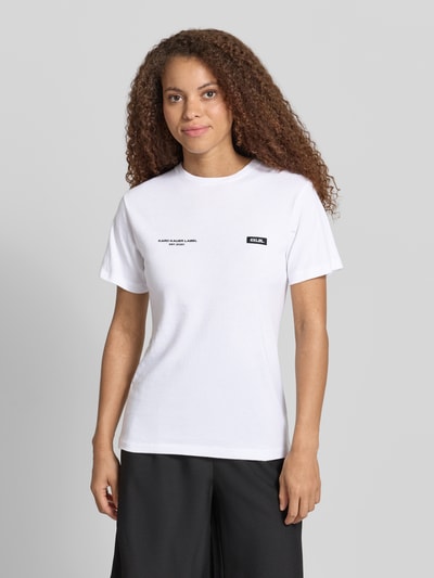 Modelka kobieca ma na sobie T-Shirt mit Logo-Print und Label-Patch od Karo Kauer, biała bawełna, krój straight fit, okrągły dekolt, długość tyłu w rozmiarze S: 62 cm, widok całej sylwetki z przodu.