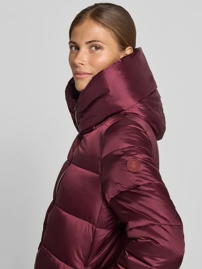 Frauenmodel trägt Steppmantel mit Kapuze Modell 'LYSA' von Save The Duck in glänzendem Dunkelrot, seitliche Eingrifftaschen, Nahaufnahme Oberkörper.