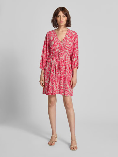 Frauenmodel trägt Minikleid aus reiner Viskose mit floralem Allover-Print von GAP, tailliert, V-Ausschnitt, 3/4-Arm, leuchtende Rottöne, Ganzkörperansicht von vorne.