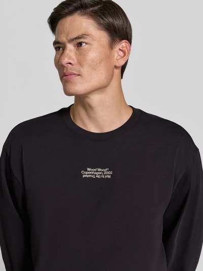 Männermodel trägt Regular Fit Longsleeve aus reiner Baumwolle Modell 'Ryan' von Wood Wood in Schwarz mit Logo-Stitching, Nahaufnahme Oberkörper. Herren Longsleeve, Rückenlänge bei Größe M 79 cm.