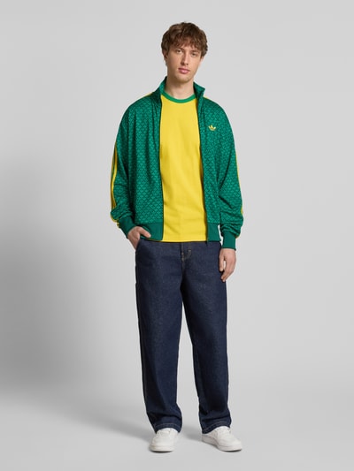 Männermodel trägt Slim Fit Jacke mit Stehkragen Modell 'Firebird' von adidas Originals in Grün mit gelben Details, Logo-Stitching, Ganzkörperansicht von vorne.