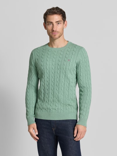 Männermodel trägt Regular Fit Pullover aus reiner Baumwolle von GANT in Hellgrün mit Zopfmuster, kombiniert mit dunkler Jeans, Ganzkörperansicht von vorne.