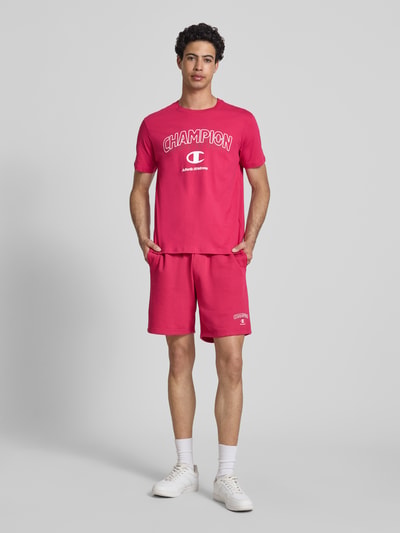 Männermodel trägt T-Shirt mit Label-Print und Rundhalsausschnitt von CHAMPION in Rot, reine Baumwolle, Regular Fit, Ganzkörperansicht von vorne.