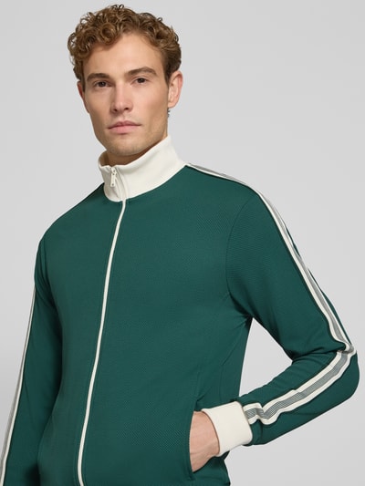 Männermodel trägt Slim Fit Sweatjacke mit Strukturmuster von MCNEAL in Dunkelgrün mit weißen Details, Reißverschluss, Nahaufnahme Oberkörper. Herren Sweatjacke, elastischer Material-Mix, Stehkragen, Eingrifftaschen, elastische Abschlüsse, Rückenlänge 68 cm, Ärmellänge 64 cm.