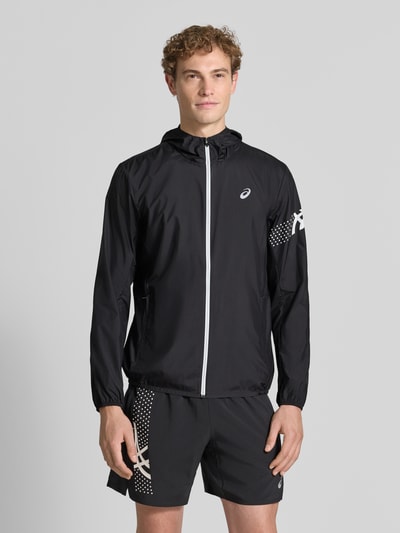 Męski model nosi Regular Fit kurtkę z nadrukiem z logo Model 'ICON' od ASICS TIGER, czarna z białym zamkiem, krój regular fit, kaptur ze stójką, elastyczne wykończenia, Długość tyłu w rozmiarze M: 72 cm, Długość rękawów w rozmiarze M: 67 cm, widok całej sylwetki z przodu.