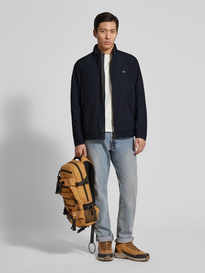 Männermodel trägt Blouson mit Logo-Print und Stehkragen von camel active in Dunkelblau, kombiniert mit Jeans und Boots, Ganzkörperansicht von vorne. Gerader Schnitt, Reißverschluss, Label-Patch, seitliche Reißverschlusstaschen, strukturiertes Material, dezenter Glanz.