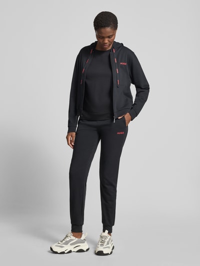 Frauenmodel trägt schwarze Regular Fit Jogginghose aus Baumwoll-Mix von HUGO mit Label Print, seitlichen Taschen und elastischem Bund, Ganzkörperansicht von vorne.