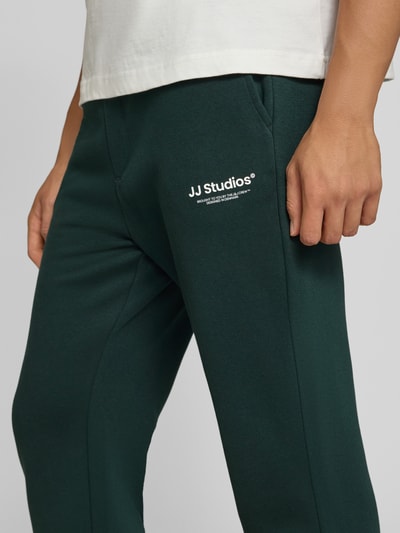Männermodel trägt Straight Fit Sweatpants aus Baumwoll-Mix Modell 'Kane' von JACK & JONES in Dunkelgrün, Logo-Detail, Nahaufnahme Oberschenkel, Herren Sweatpants, elastischer Bund, Gesäßtasche, elastische Beinabschlüsse, Innenbeinlänge 72 cm bei Größe M