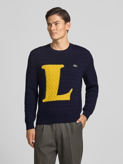 Männermodel trägt Regular Fit Strickpullover aus Baumwoll-Mix von Lacoste, dunkelblau mit gelbem L, Grobstrick, Ganzkörperansicht von vorne. Herren Strickjacke von Lacoste