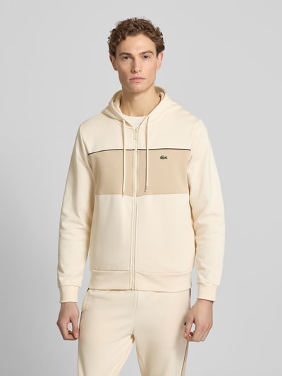 Männermodel trägt Regular Fit Sweatjacke aus reiner Baumwolle von Lacoste in Beige mit Kontraststreifen, Kapuze, Logo-Patch, Ganzkörperansicht von vorne.