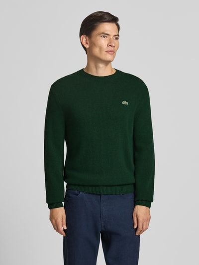 Männermodel trägt dunkelgrünen Regular Fit Pullover aus reiner Wolle von Lacoste, kombiniert mit dunkelblauer Hose, Ganzkörperansicht von vorne. Herren Pullover von Lacoste.