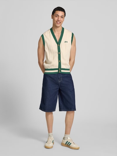 Mannenmodel draagt Regular Fit Weste in Strick-Optik mit Logo-Stitching van BLKVIS in crème en groen met V-hals, knopen, opgestikte zakken, ganzekörperaanzicht von vorne.