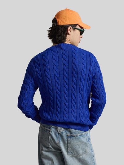 Männermodel trägt Regular Fit Strickpullover aus reiner Baumwolle von Polo Ralph Lauren w kolorze niebieskim, ścieg warkoczowy, krój regular fit, imitacja dzianiny, detal z logo marki, długi rękaw, prążkowane wykończenie, dzianina z bawełny, widok całej sylwetki z tyłu.