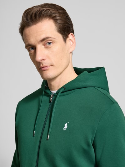 Mannelijk model draagt Regular Fit Sweatjacke aus Baumwoll-Mix van Polo Ralph Lauren in effen donkergroen met logo-stiksel en capuchon, Nahaufnahme bovenlichaam.