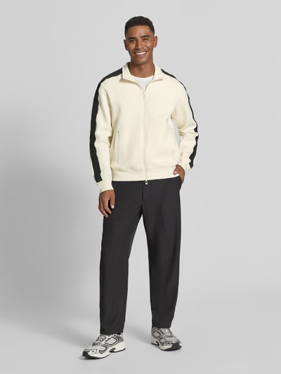 Männermodel trägt Regular Fit Sweatjacke aus Baumwoll-Mix Modell 'Felpa' von Armani Exchange in Creme mit schwarzen Details, Ganzkörperansicht von vorne, Herren Sweatjacke mit Stehkragen, Zweiwege-Reißverschluss und elastischen Abschlüssen.