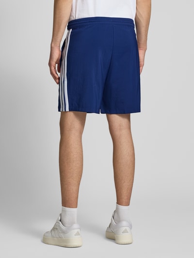 Männermodel trägt Regular Fit Sport-Shorts mit elastischem Bund von adidas Sportswear in Blau mit weißen Kontraststreifen, Ganzkörperansicht Rückansicht.