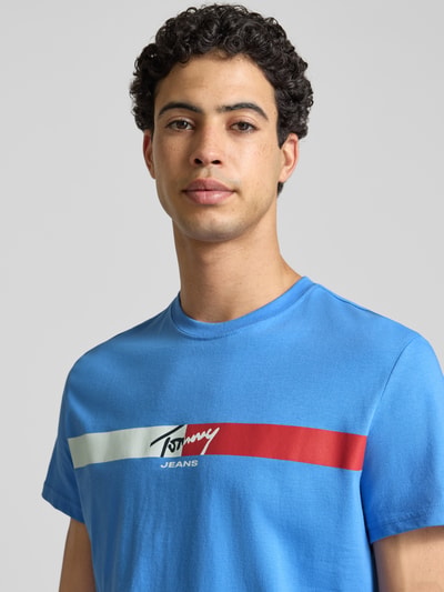 Männermodel trägt Regular Fit T-Shirt aus reiner Baumwolle von Tommy Jeans in Blau mit Label Print, Jersey-Textur, Nahaufnahme Oberkörper.