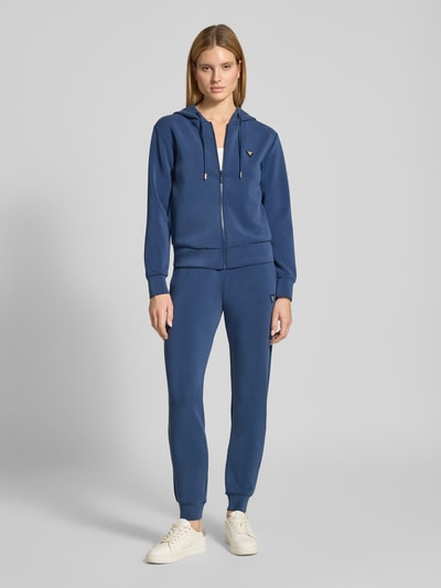 Vrouwenmodel draagt Regular Fit Sweatshirt met logo-applicatie model 'Olympe' van Guess Activewear, elastische materiaalmix met modal, capuchon met trekkoord, rits, elastische boorden, blauw, effen ontwerp, gladde textuur, volledige lichaamsweergave van voren.
