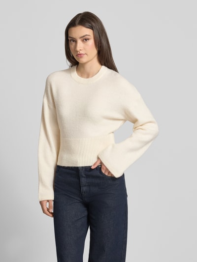 Frauenmodel trägt Strickpullover mit weiten Ärmeln Modell 'Stella' von Gina Tricot im Material-Mix mit Alpaka-Anteil, Regular-Fit, Rundhalsausschnitt, weiche cremefarbene Textur, Damen Strickpullover, Rückenlänge 55 cm, Ärmellänge 60 cm, Ganzkörperansicht von vorne.
