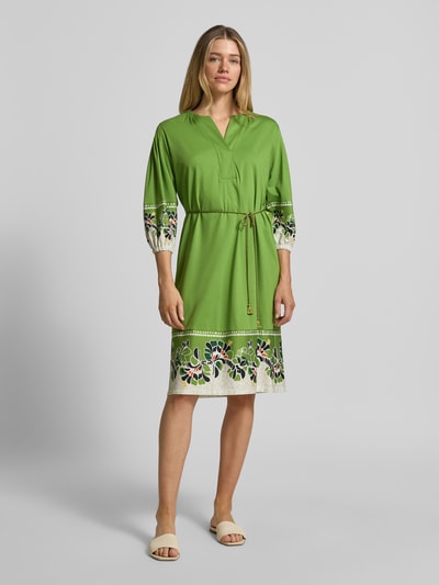 Frauenmodel trägt Knielanges Kleid mit Bindegürtel von Marc Cain in Grün mit bunten Stickereien, reine Baumwolle, gerader Schnitt, V-Ausschnitt, Bindegürtel, knielang, Ganzkörperansicht von vorne.