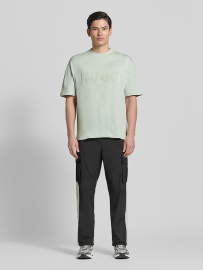 Männermodel trägt Relaxed Fit T-Shirt aus reiner Baumwolle Modell 'DINKEE' von HUGO in Optik: Unifarben, Passform: Relaxed-Fit, Applikation: 3D Label-Print, Rundhals-Ausschnitt, Ärmelform: Kurzarm, Materialverarbeitung: Jersey, Ganzkörperansicht von vorne.